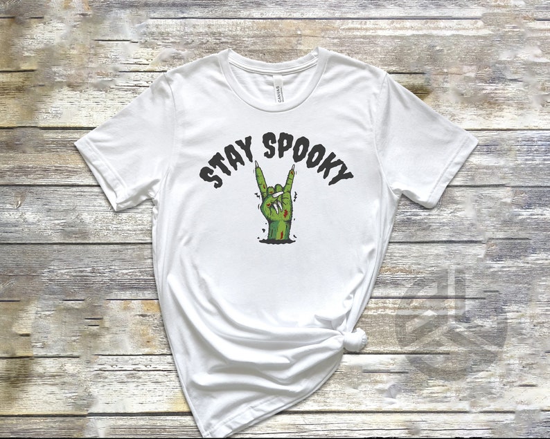 Puede incluir: Camiseta blanca con una mano de zombie verde haciendo el signo de rock and roll. El texto "Stay Spooky" est&aacute; por encima de la mano.
