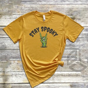 Puede incluir: Una camiseta amarilla mostaza con el texto "Stay Spooky" y una mano de zombie verde haciendo un gesto de rock and roll.