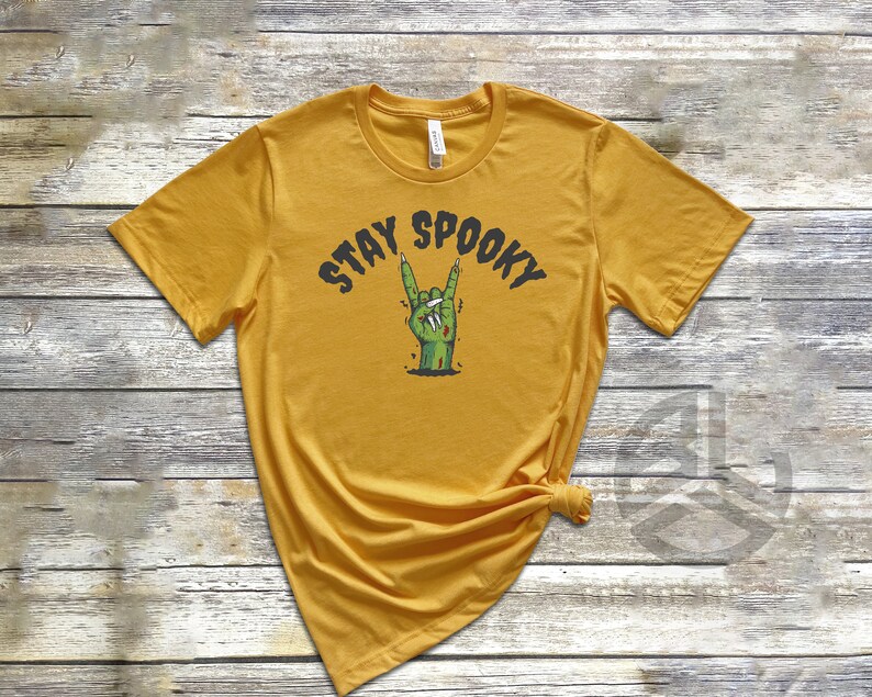 Puede incluir: Una camiseta amarilla mostaza con el texto "Stay Spooky" y una mano de zombie verde haciendo un gesto de rock and roll.