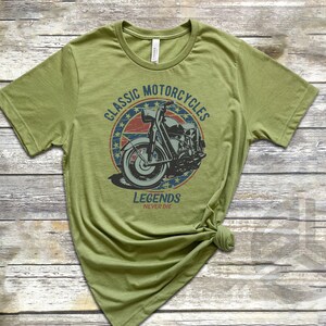 Op de afbeelding: Een groen T-shirt met een vintage-stijl afbeelding van een motorfiets. De afbeelding heeft een rood, wit en blauw kleurenschema en de tekst "Classic Motorcycles Legends Never Die".