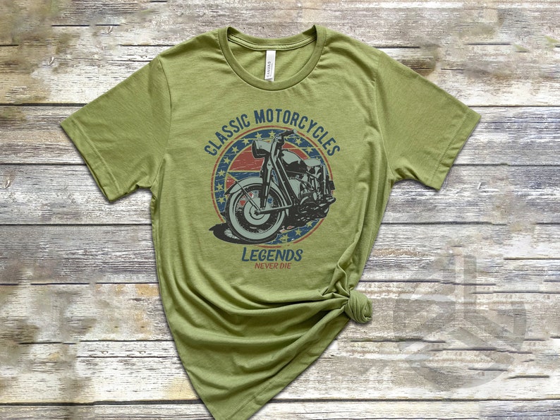 Op de afbeelding: Een groen T-shirt met een vintage-stijl afbeelding van een motorfiets. De afbeelding heeft een rood, wit en blauw kleurenschema en de tekst "Classic Motorcycles Legends Never Die".