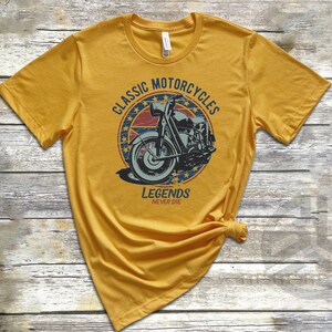 Op de afbeelding: Een geel T-shirt met een vintage-stijl grafische afbeelding van een motorfiets. De afbeelding bevindt zich in een cirkel met de tekst "Classic Motorcycles" en "Legends Never Die".