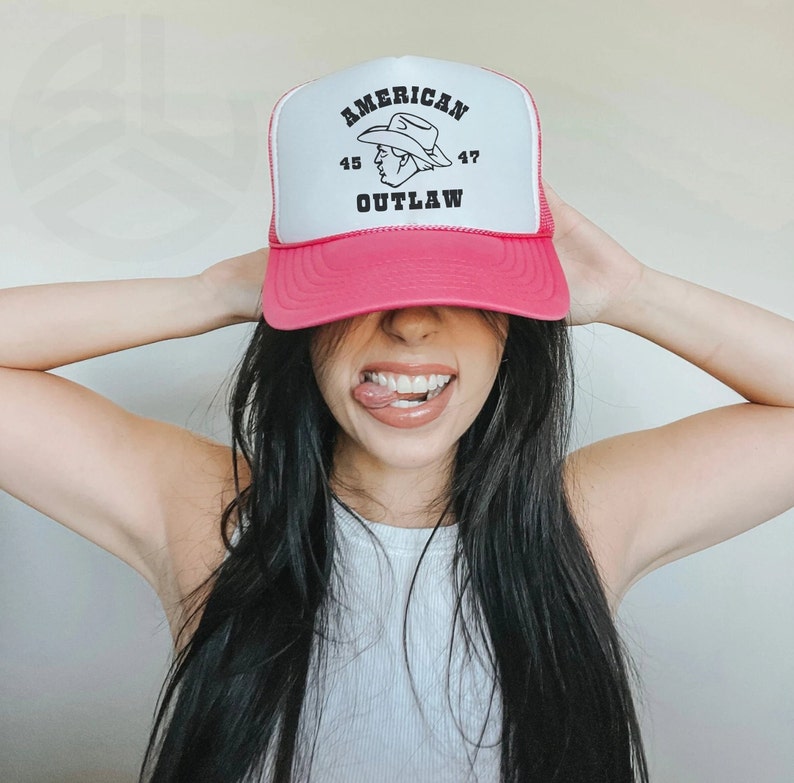May include: A pink trucker hat with a white logo that says "AMERICAN OUTLAW" and a cartoon image of a cowboy wearing a hat. The numbers "45" and "47" are on either side of the logo.