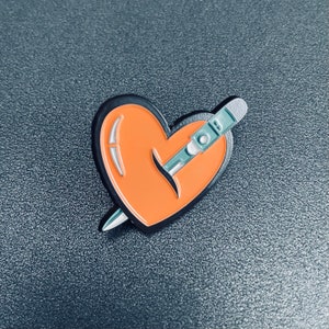 May include: A black and orange enamel pin shaped like a heart with a silver needle piercing through it.