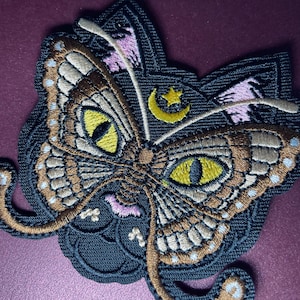 May include: Embroidered patch featuring a moth design with intricate details. The moth has large yellow eyes, brown and tan wings, and a crescent moon and star. The patch is on a dark background with pink accents.
