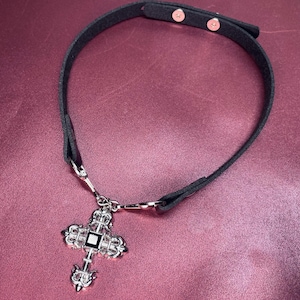 May include: Black choker necklace with a silver cross pendant. The cross has ornate detailing and a central square accent. The necklace features a black strap with adjustable snap closures. The pendant is approximately 2 inches in length.
