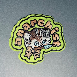 May include: Embroidered patch featuring a cartoon kitten with brown and white fur, blue eyes, and a bow. The kitten is holding a small blue object. The patch has the word "march is" in orange letters on a green background.