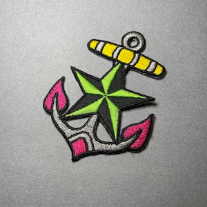 May include: Embroidered anchor patch featuring a black and green star, yellow crossbar, and pink flame-like accents. The anchor is outlined in gray and is a nautical-themed accessory.