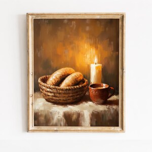 May include: An oil painting in a gold frame depicting a woven basket filled with two oval-shaped loaves of bread, a brown ceramic mug, and a lit white candle, all set against a warm, textured brown background with soft golden light.