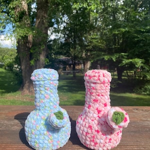 Op de afbeelding: Twee gehaakte waterpijpen gemaakt van zacht, dik garen. De ene pijp heeft een lichtblauwe basis met roze en witte spikkels, terwijl de andere een lichtroze basis heeft met donkerroze en witte spikkels. Beide hebben een klein groen bladaccent.