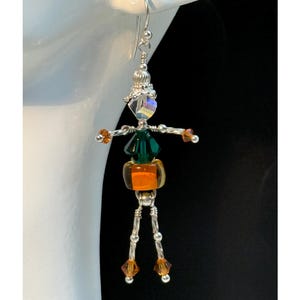 May include: A dangle earring shaped like a human figure, constructed from silver-tone wire and beads. The head is a clear iridescent crystal, the torso is a dark green crystal, and the skirt is an orange glass bead. Arms and legs feature small orange crystal accents.