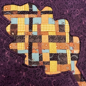 May include: A quilted leaf-shaped design with a patchwork center. The leaf is outlined in purple fabric with a floral pattern, and the center features a mix of orange, yellow, blue, and brown fabrics. The background has a floral pattern.