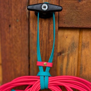 May include: A long, bright neon pink leash with a teal strap handle and accents. The leash features gold-toned metal swivel snap hooks and rivets, with a coiled design hanging from a black T-shaped handle mounted on a wooden surface.