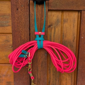 May include: A bright pink dog leash is coiled and hanging on a wooden door. The leash is accented with teal leather straps and gold-colored metal hardware. The leash is attached to a black handle.