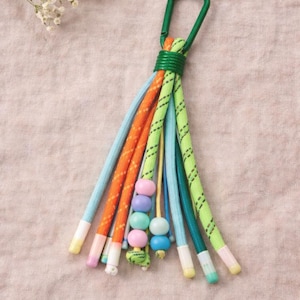 May include: A colorful keychain with a green carabiner clip. The keychain features multiple cords in various colors, including orange, blue, and green, with decorative beads. The cords are approximately 6 inches long.