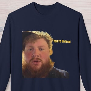 May include: Navy blue sweatshirt featuring a meme of a man with a beard and the text "You're Banned" in yellow. The image is centered on the front of the sweatshirt, creating a humorous and eye-catching design.