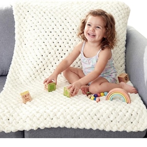 Peut inclure: Une couverture en tricot épais de couleur crème avec une texture tissée. Un jeune enfant est assis sur la couverture, jouant avec des blocs de bois et un jouet en forme d'arc-en-ciel. La couverture est posée sur un canapé gris.