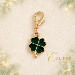 May include: A gold-toned metal charm featuring a four-leaf clover design with deep green enamel inlay. The clover is attached to a gold-toned lobster claw clasp by a small circular jump ring. The charm has a polished finish and a smooth surface.