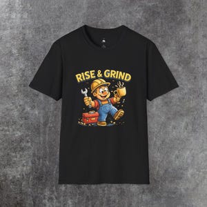 Puede incluir: Camiseta negra con un dibujo animado de un trabajador de la construcción sosteniendo una llave inglesa y una taza de café. El diseño incluye una caja de herramientas y las palabras "RISE & GRIND" en amarillo. El trabajador lleva un casco amarillo y un mono azul.