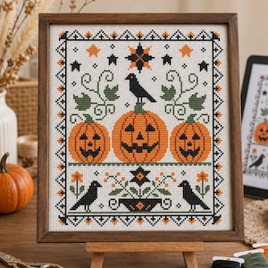 May include: A framed cross-stitch embroidery featuring three orange jack-o'-lanterns, black crows, and stars. The design includes green vines and a geometric border in black and orange, all displayed within a dark wooden frame on a small stand.
