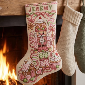 May include: A cream-colored Christmas stocking featuring a cross-stitched design of a nutcracker, a gingerbread house, a cupcake, and a lollipop. The name JEREMY is stitched in green at the top. The stocking is decorated with pink and green accents.
