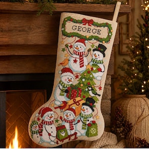 May include: A beige Christmas stocking featuring a cross-stitch design of multiple snowmen, a decorated pine tree, and wrapped gifts. The name GEORGE is stitched in black letters inside a green wreath at the top. The stocking has a hanging loop.