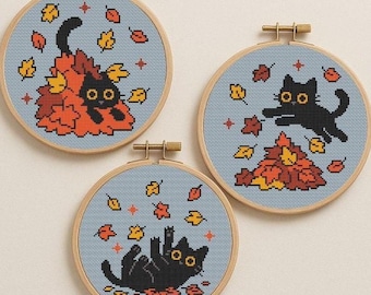 May include: Three cross-stitch hoops featuring black cats playing in autumn leaves. The cats are surrounded by orange, yellow, and red leaves on a light blue background. The hoops are framed in natural wood.