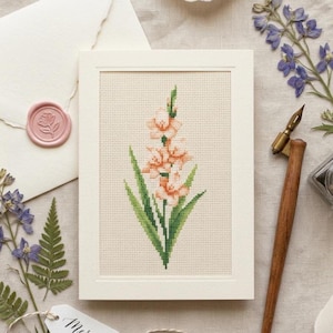 May include: A framed cross-stitch of a peach-colored gladiolus flower with green leaves. The artwork is in a white frame, with a pink wax seal, dried flowers, and a calligraphy pen nearby. The overall theme is floral and handmade.