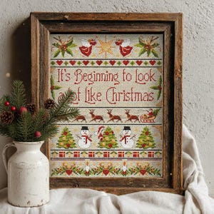 May include: A framed cross-stitch artwork in a wooden frame. The artwork displays the text "It's Beginning to look a lot like Christmas" in red, alongside festive images of reindeer, snowmen, and Christmas trees.