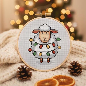 May include: A cross-stitch embroidery hoop showing a sheep decorated with festive Christmas lights. The sheep has a white body, tan face and ears, and is framed by a wooden hoop. The background includes a soft, white blanket, pine cones, and dried orange slices.