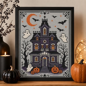 May include: A framed cross-stitch embroidery featuring a dark purple haunted house with glowing orange windows, surrounded by white ghosts, black bats, and orange pumpkins under a crescent moon, all set against a light grey background.