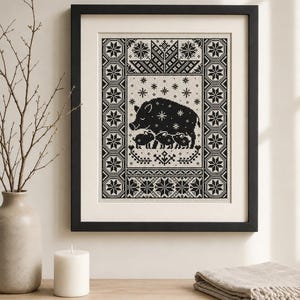 Può includere: Un ricamo a punto croce incorniciato che presenta un disegno in bianco e nero di un grande cinghiale con tre cuccioli. Il motivo centrale è circondato da motivi geometrici a fiocco di neve ed elementi floreali, il tutto in una cornice nera.