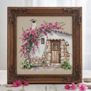 Könnte beinhalten: Kreuzstich-Kunstwerk eines malerischen Häuschens mit Steinfassade, Holztür und leuchtend rosa Bougainvillea. Das Kunstwerk ist in einem detaillierten, verzierten braunen Holzrahmen gerahmt, was eine rustikale und charmante Ästhetik erzeugt.