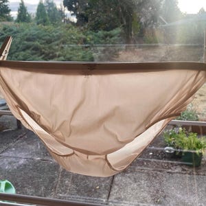 Könnte beinhalten: Ein beigefarbenes Unterwäschestück hängt an einer Wäscheleine, befestigt mit Holzwäscheklammern. Der Stoff ist glatt und scheint aus einem leichten Material zu bestehen. Der Hintergrund zeigt eine verschwommene Außenszene mit Grün.