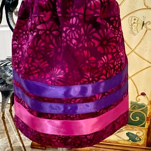 May include: A vibrant, handmade skirt featuring a floral pattern in shades of purple and pink. The skirt is adorned with horizontal bands of satin ribbon in purple and pink. The fabric has a textured appearance, and the skirt is gathered at the waist.