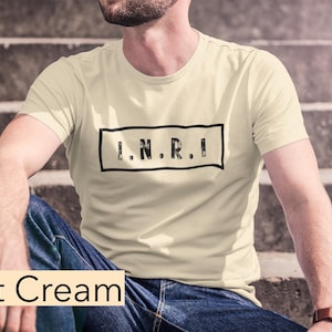 May include: A soft cream colored t-shirt with a black rectangle containing the text "I.N.R.I." in black.