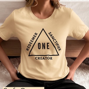 Könnte beinhalten: Ein T-Shirt in zartem Creme mit einem schwarzen Dreiecks-Motiv. Das Dreieck hat die Worte "Redeemer", "Sanctifier", "Creator" und "ONE" aufgedruckt.