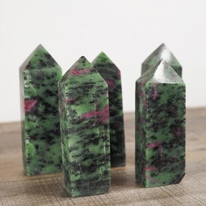 May include: Five green and pink gemstone points, also known as zoisite, are arranged on a wooden surface. The points are polished and have a natural, irregular shape.