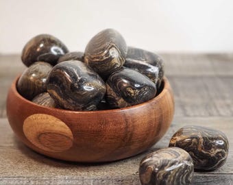 May include: A wooden bowl filled with polished stones. The stones are a mix of dark brown and black with swirling patterns. Two stones rest outside the bowl on a wooden surface. The bowl is made of a light brown wood.