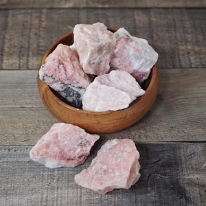 May include: A wooden bowl filled with raw, pink rose quartz crystals. The crystals vary in size and shape, with some showing streaks of darker pink and gray. The bowl sits on a weathered wooden surface, with a few crystals scattered around it.