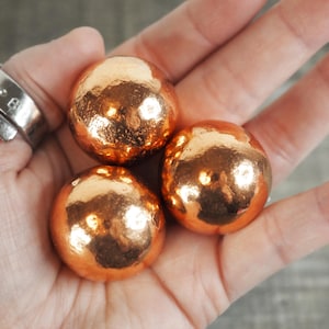 May include: Three copper-colored spheres with a textured, metallic finish are held in a hand. The spheres have a reflective surface, catching the light and creating highlights. The overall effect is one of warmth and richness.