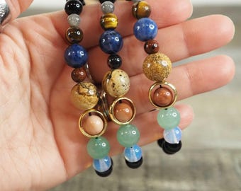 May include: Three beaded bracelets featuring various coloured stones, including brown, blue, green, and gold. The bracelets are layered on top of each other and are being held by a hand.