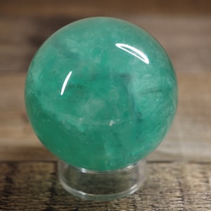 May include: A smooth, polished green fluorite sphere on a clear stand. The sphere has a translucent, cloudy appearance with swirls of color.
