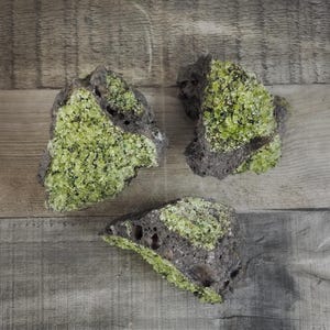 May include: Three rough, dark gray rocks with vibrant, lime green crystal formations. The rocks are irregularly shaped and have a porous texture, set against a light gray wood grain background. The crystals add a striking contrast.