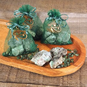 May include: Three green organza bags filled with green gemstones and gold charms with the Eye of Horus symbol. The bags are on a wooden tray with loose green gemstones scattered around them.
