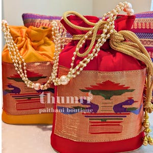 May include: Two drawstring bags, one red and one yellow, with gold and pearl handles. The bags feature a decorative panel with a colorful peacock design and the text "thumri paithani boutique".