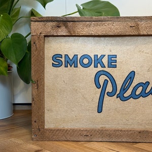 Puede incluir: Un letrero de madera con marco marr&oacute;n y fondo beige. El letrero dice "SMOKE PLANTS" en letras azules.
