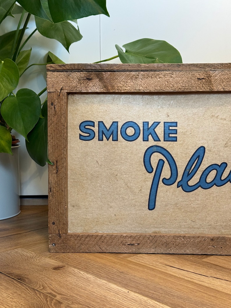 Puede incluir: Un letrero de madera con marco marr&oacute;n y fondo beige. El letrero dice "SMOKE PLANTS" en letras azules.