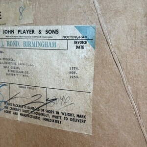 Puede incluir: Una caja de cart&oacute;n marr&oacute;n vintage con una etiqueta azul y blanca. La etiqueta dice "John Player & Sons" y "J. Bond, Birmingham". La etiqueta tambi&eacute;n incluye una fecha de factura del "13 de noviembre de 1950". La caja tiene una nota manuscrita que dice "Peso bruto 2040".
