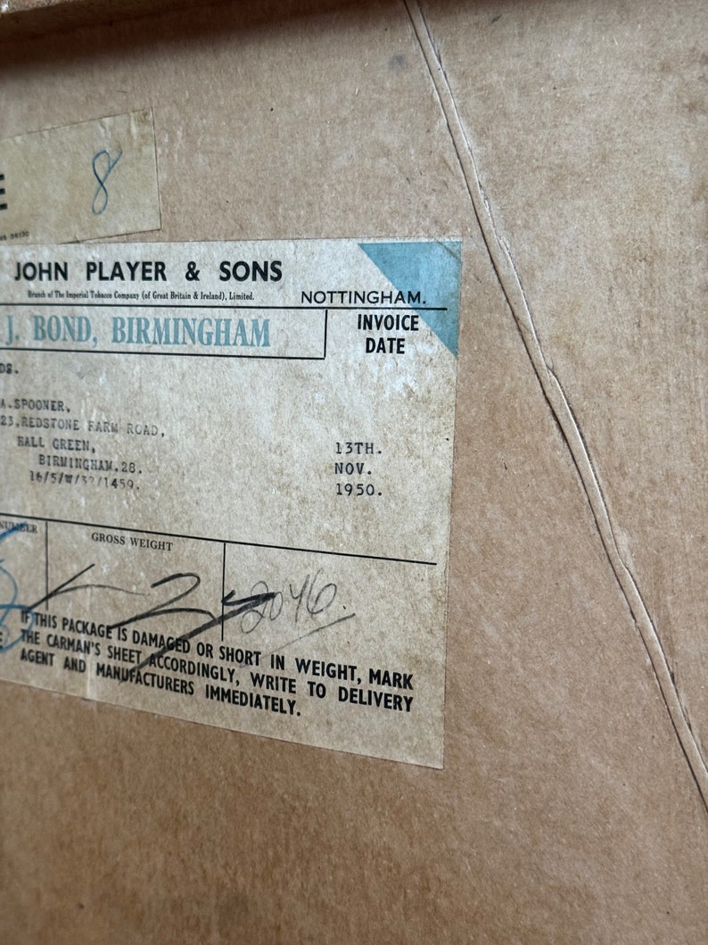 Puede incluir: Una caja de cart&oacute;n marr&oacute;n vintage con una etiqueta azul y blanca. La etiqueta dice "John Player & Sons" y "J. Bond, Birmingham". La etiqueta tambi&eacute;n incluye una fecha de factura del "13 de noviembre de 1950". La caja tiene una nota manuscrita que dice "Peso bruto 2040".